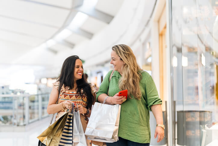 Two women retail shopping in a mall, holding shopping bags, and smiling at one another.
